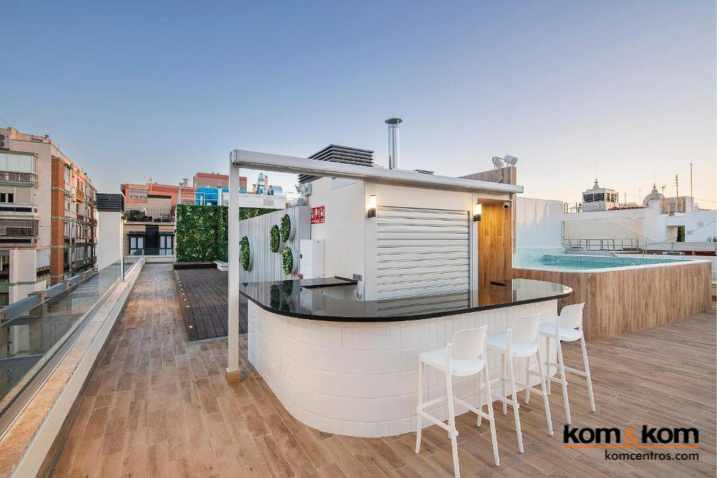 Bar de terraza en azotea de hotel con encimera de acero inoxidable, estantes de cristal con botellas de c&oacute;ctel variadas, cocteleras y fregadero. Dise&ntilde;o elegante y profesional para bar de exteriores.